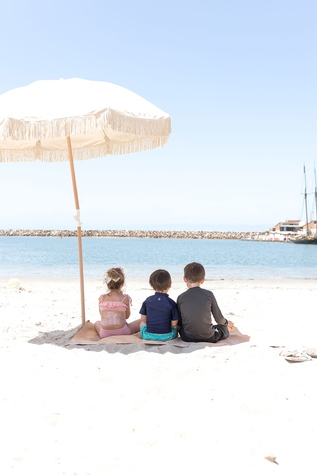 three kids sitting on the beach under an umbrella